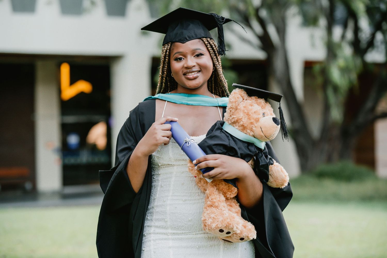 A graduate in cap and gown holds a diploma and plush teddy bear during outdoor commencement ceremony.