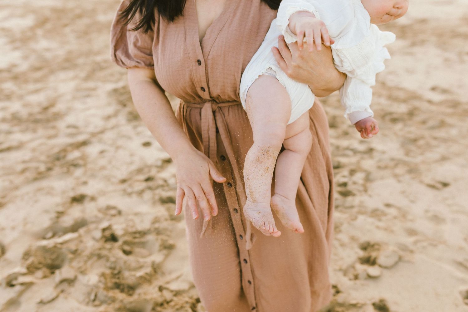 A parent in a beige coat holds a baby in white clothing while walking on a sandy beach.