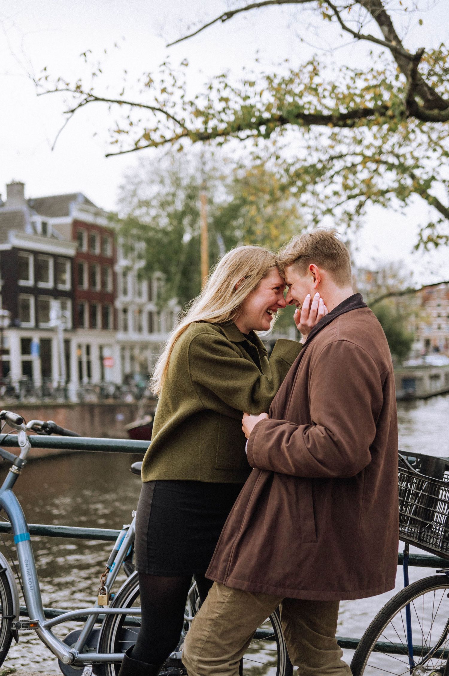 Couple standing close together by a canal in Amsterdam, romantic proposal moment with bicycles in the background.