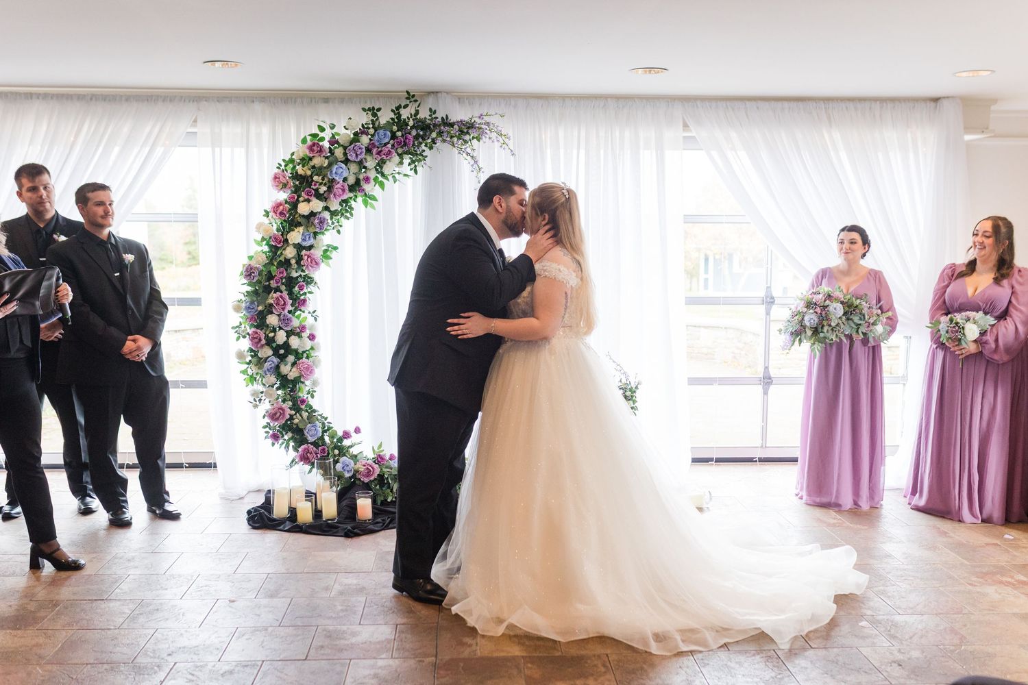 A couple shares their first kiss during a wedding ceremony under a floral arch with attendants in lavender dresses nearby.