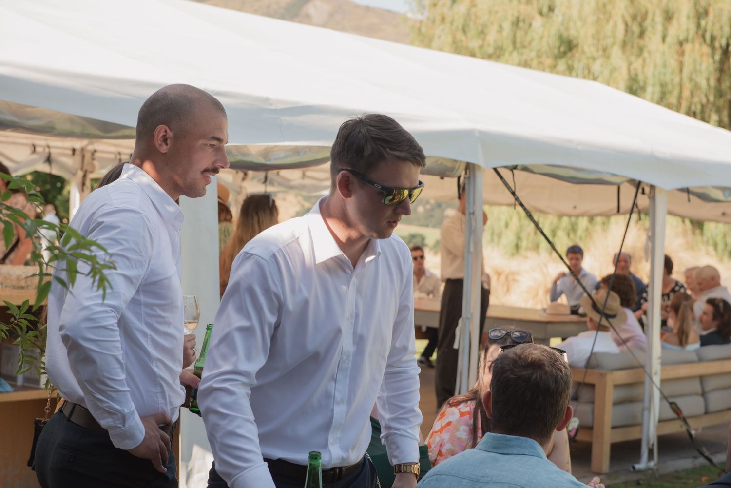 Two people in white dress shirts converse under a white tent canopy at an outdoor event.