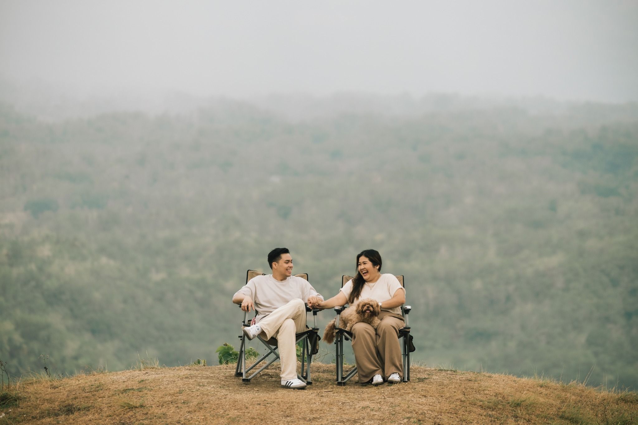 A couple sits together on a hilltop overlooking a misty mountain landscape during a romantic outdoor photo session.