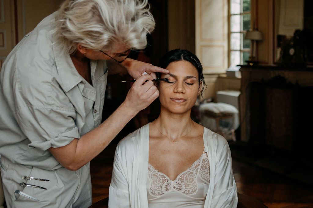 Makeup artist applying eyeshadow to a bride in a lace robe during wedding morning preparations.
