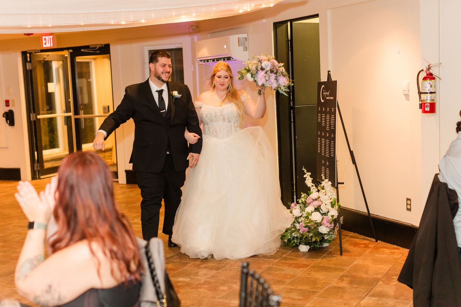Wedding party members walk through an elegant hallway with warm lighting and white walls during a reception.