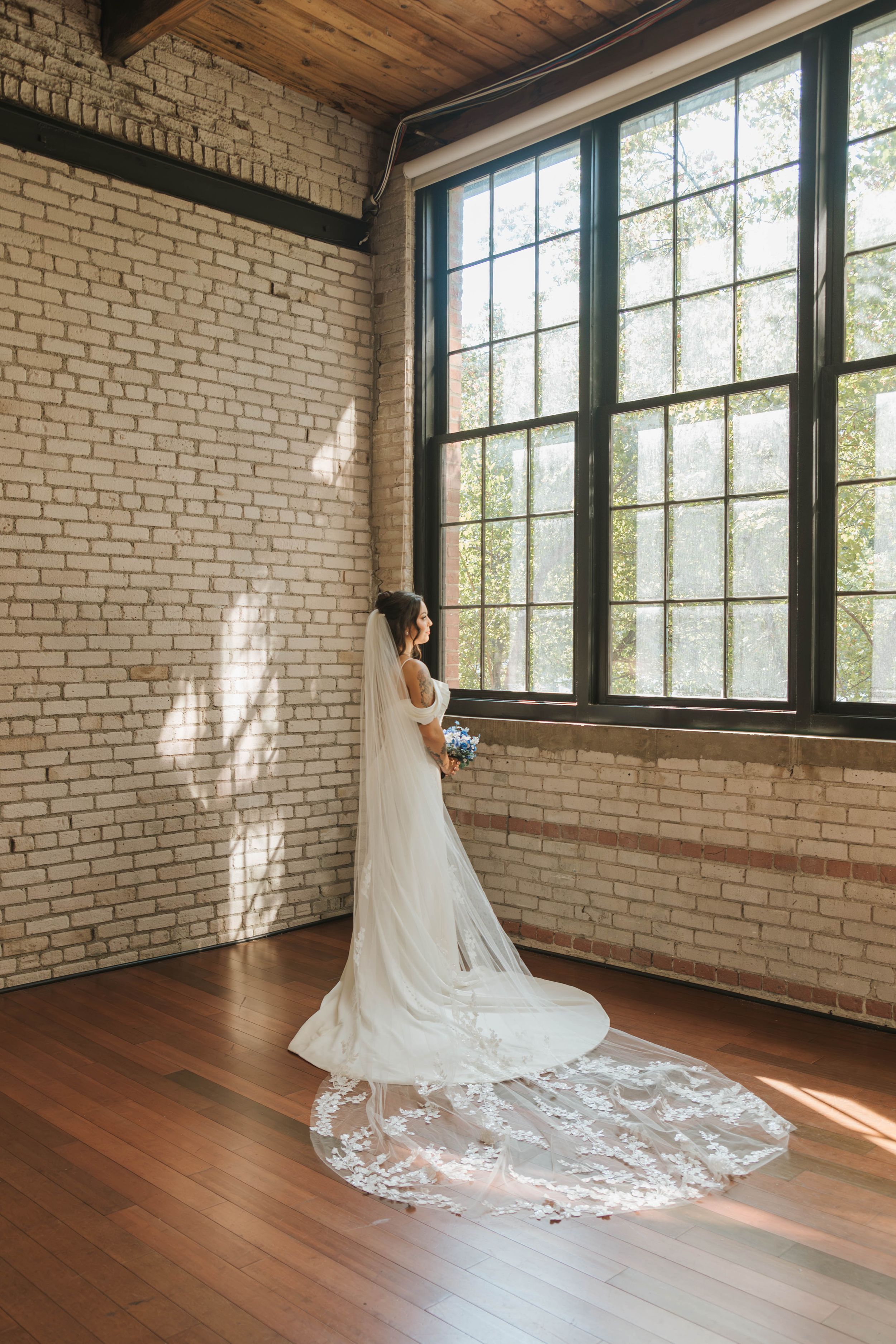 A bride in a flowing white wedding gown with a long lace train poses in a sunlit brick-walled room with hardwood floors.