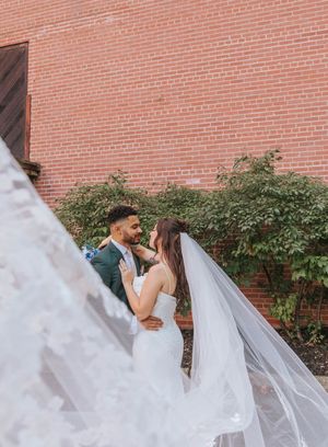 A romantic wedding couple shares a tender moment against a rustic brick wall with flowing veil and greenery.