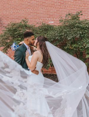 A romantic wedding couple shares a kiss as the bride's long veil flows dramatically in front of a brick wall.