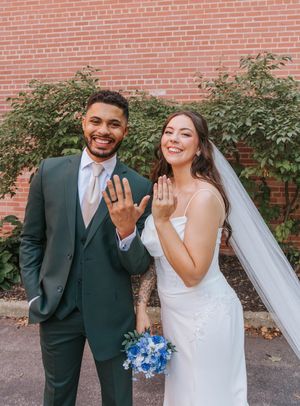 A happy couple in wedding attire poses against a brick wall with blue floral bouquet details.
