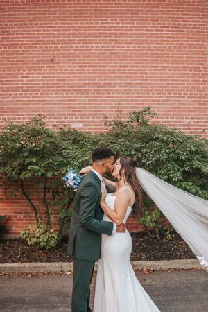 Newlyweds share a kiss against a red brick wall backdrop as the bride's veil flows in the breeze.