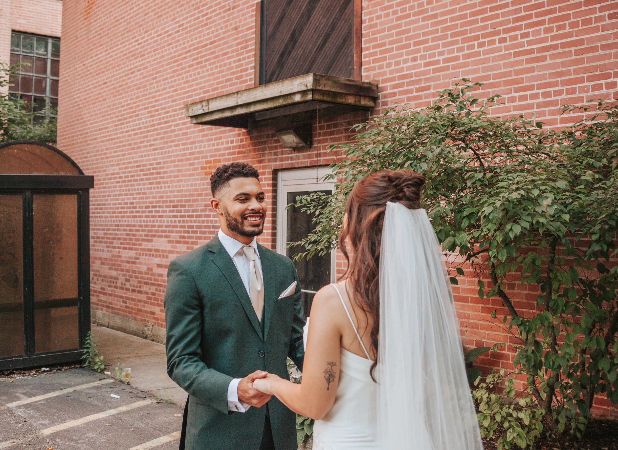 A couple in wedding attire embraces in front of a rustic brick wall with greenery.