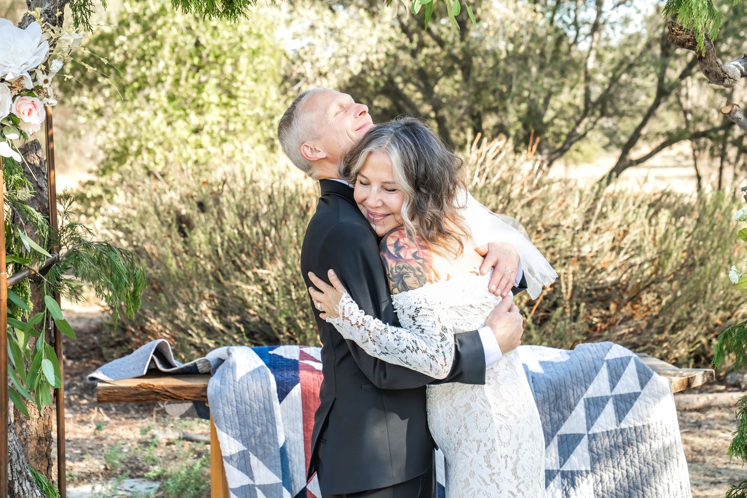 Bride and groom share a joyful embrace outdoors at a rustic woodland wedding ceremony.