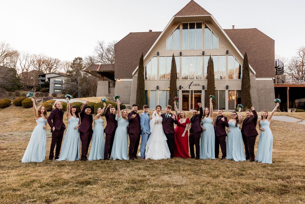 Wedding party celebrating outdoors in front of a modern chapel, bridesmaids in blue, groomsmen in burgundy, arms raised.