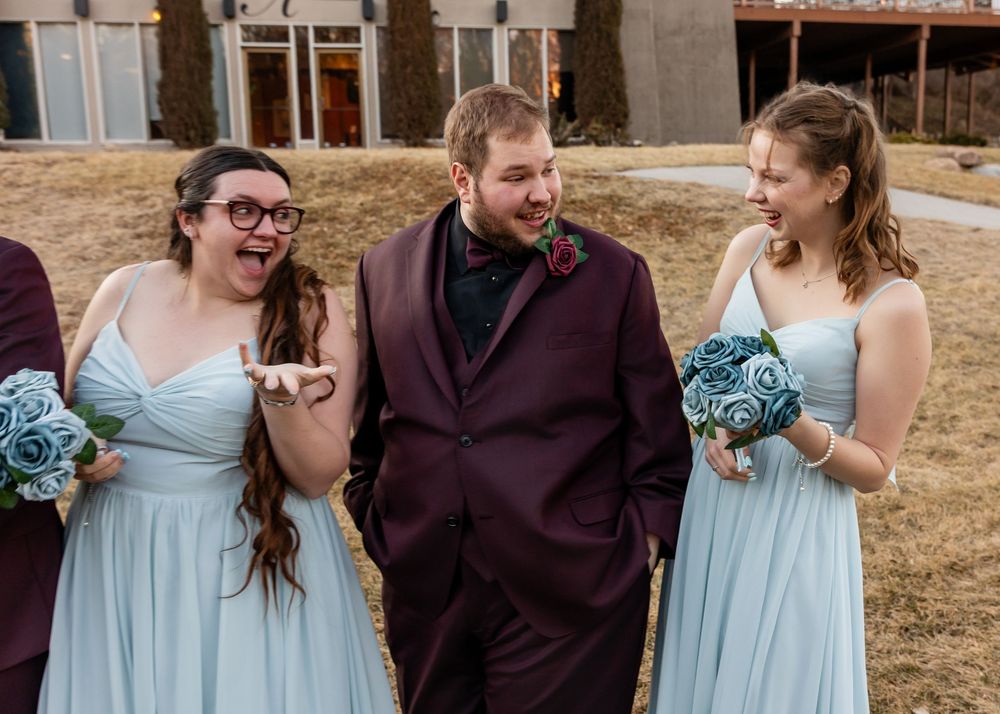 Groom in burgundy suit laughing with two bridesmaids in light blue dresses holding teal bouquets at wedding.
