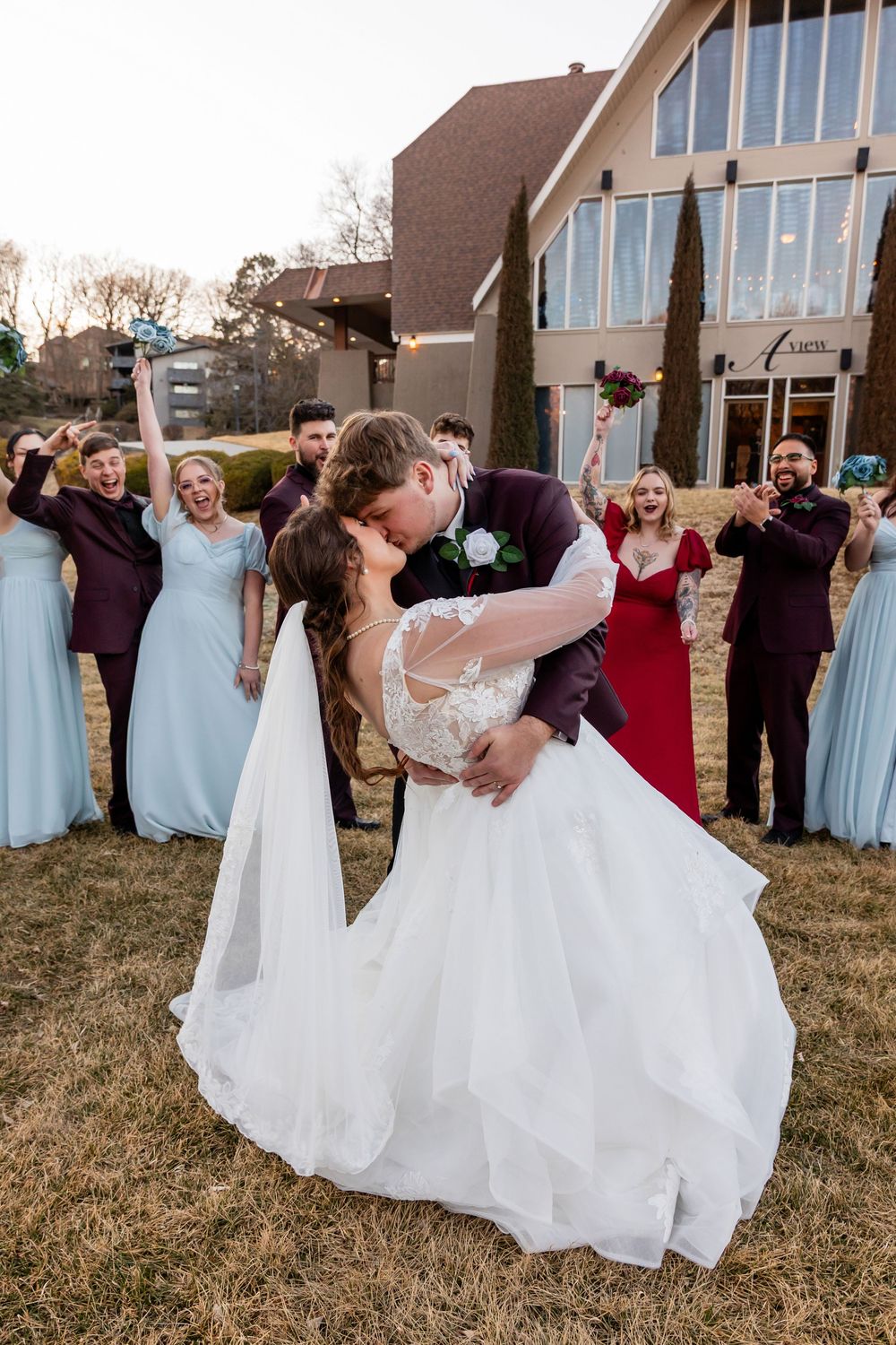 Bride and groom share a romantic dip kiss outdoors while wedding party celebrates behind them near a modern church.