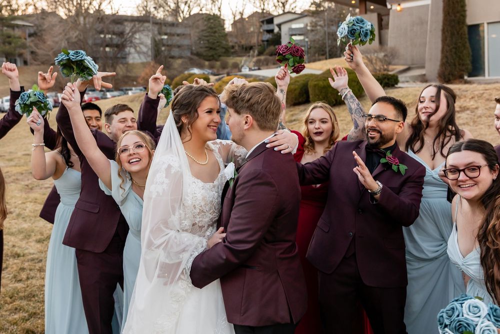 Wedding party celebrating bride and groom kiss outdoors, bridesmaids in blue and burgundy dresses raising bouquets.