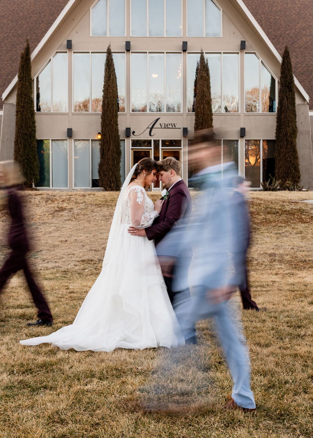 Bride and groom share a romantic moment outside a wedding venue while blurred guests walk past in the foreground.