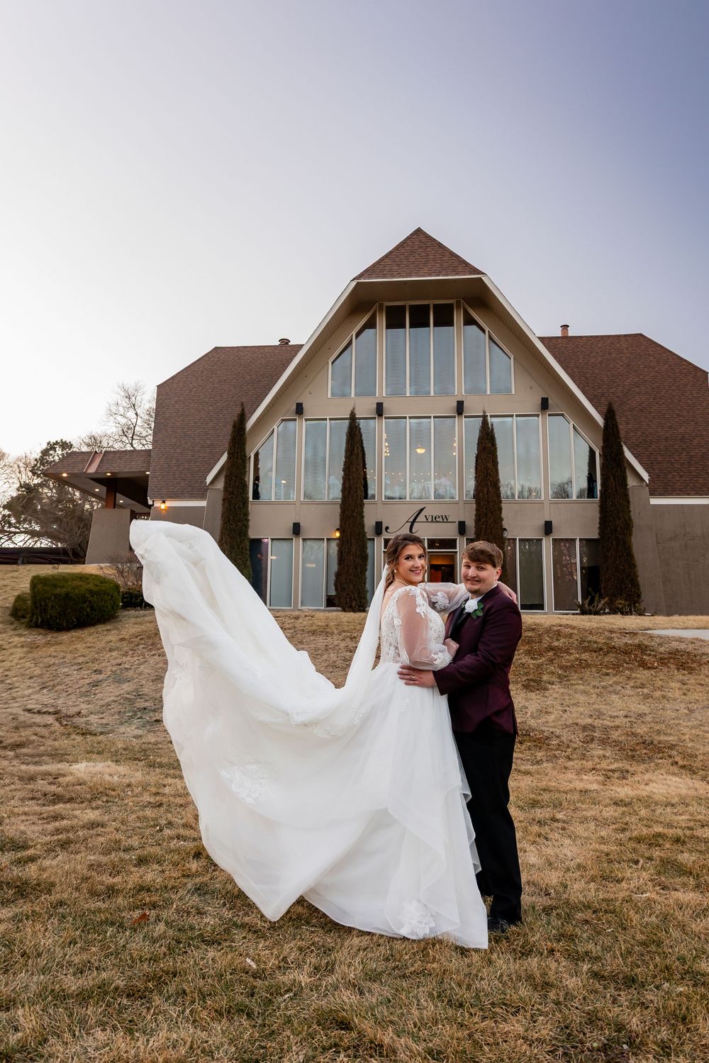 Bride and groom pose outside a modern A-frame church, her white gown flowing in the wind on their wedding day.