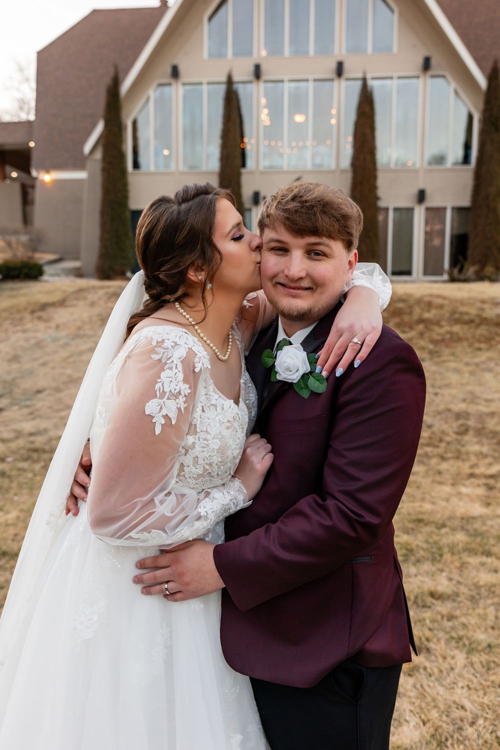 Bride in lace gown kisses groom in burgundy suit outside church on wedding day.