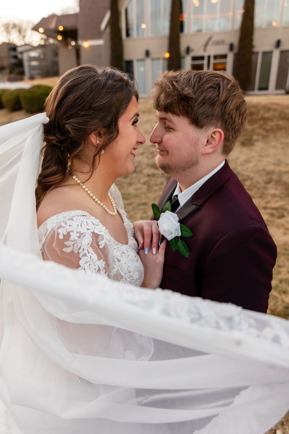 A bride and groom share a romantic moment outdoors, her veil flowing, as she wears lace and he wears a burgundy suit.