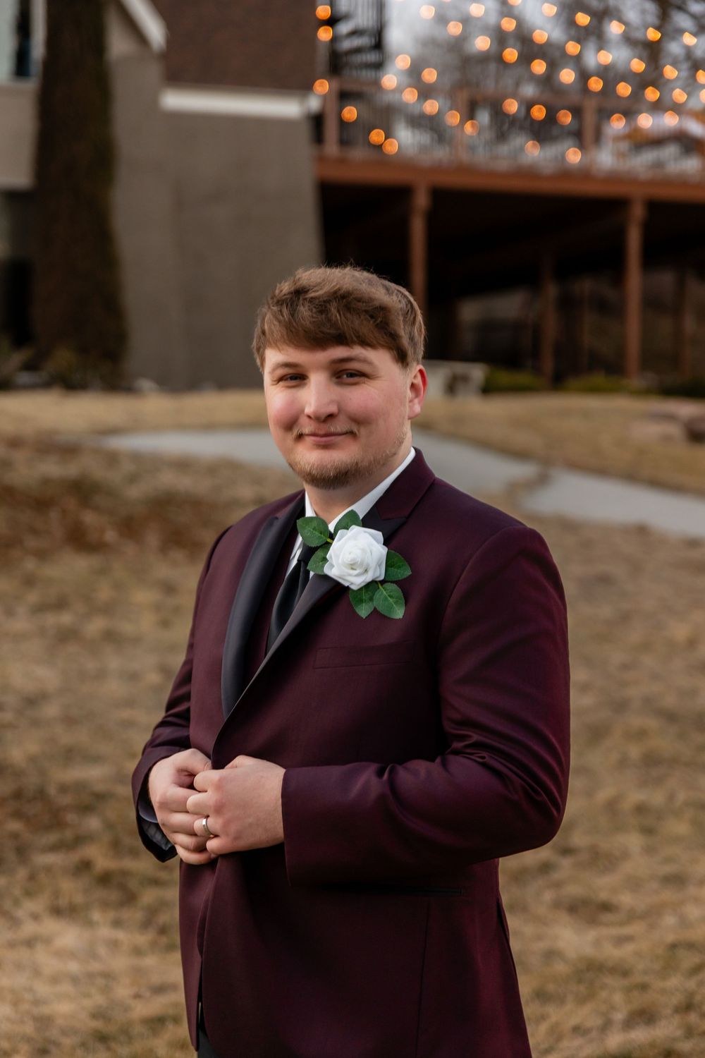 Groom in burgundy tuxedo with white rose boutonniere smiles confidently outdoors at evening wedding reception.