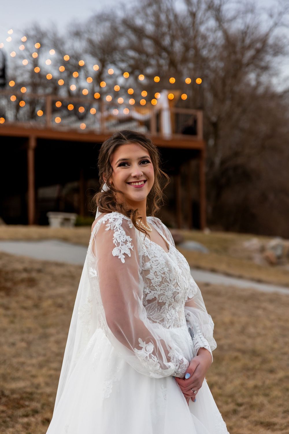 Bride in lace wedding gown with sheer sleeves smiling outdoors with string lights.
