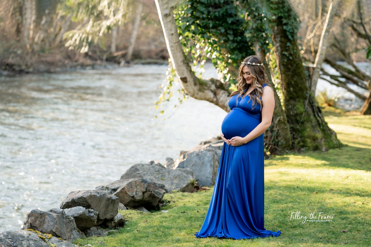 Maternity photoshoot by river with woman in flowing blue dress among rocks and trees.