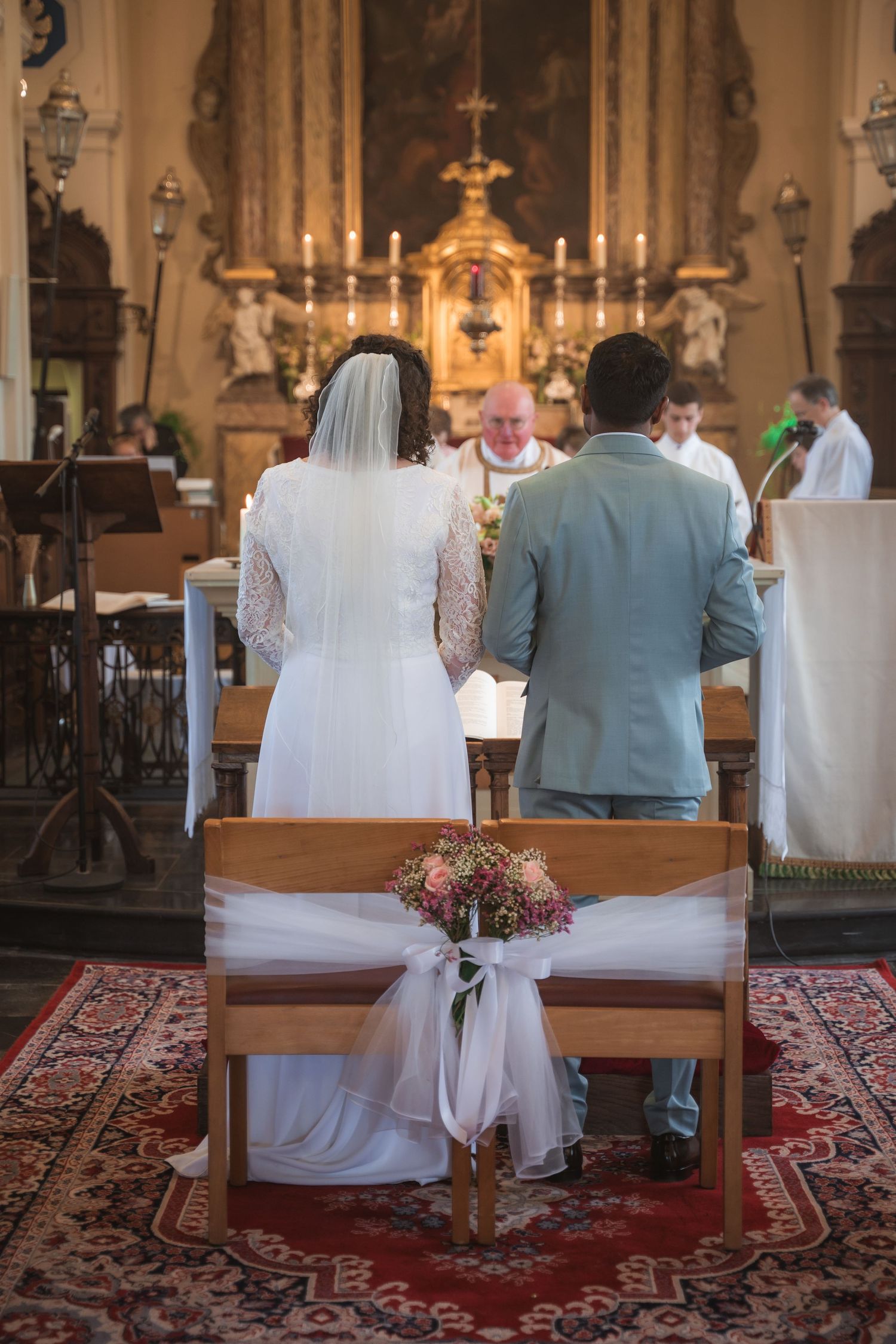 Bride and groom standing at church altar during wedding ceremony with priest and floral bouquet on pew.