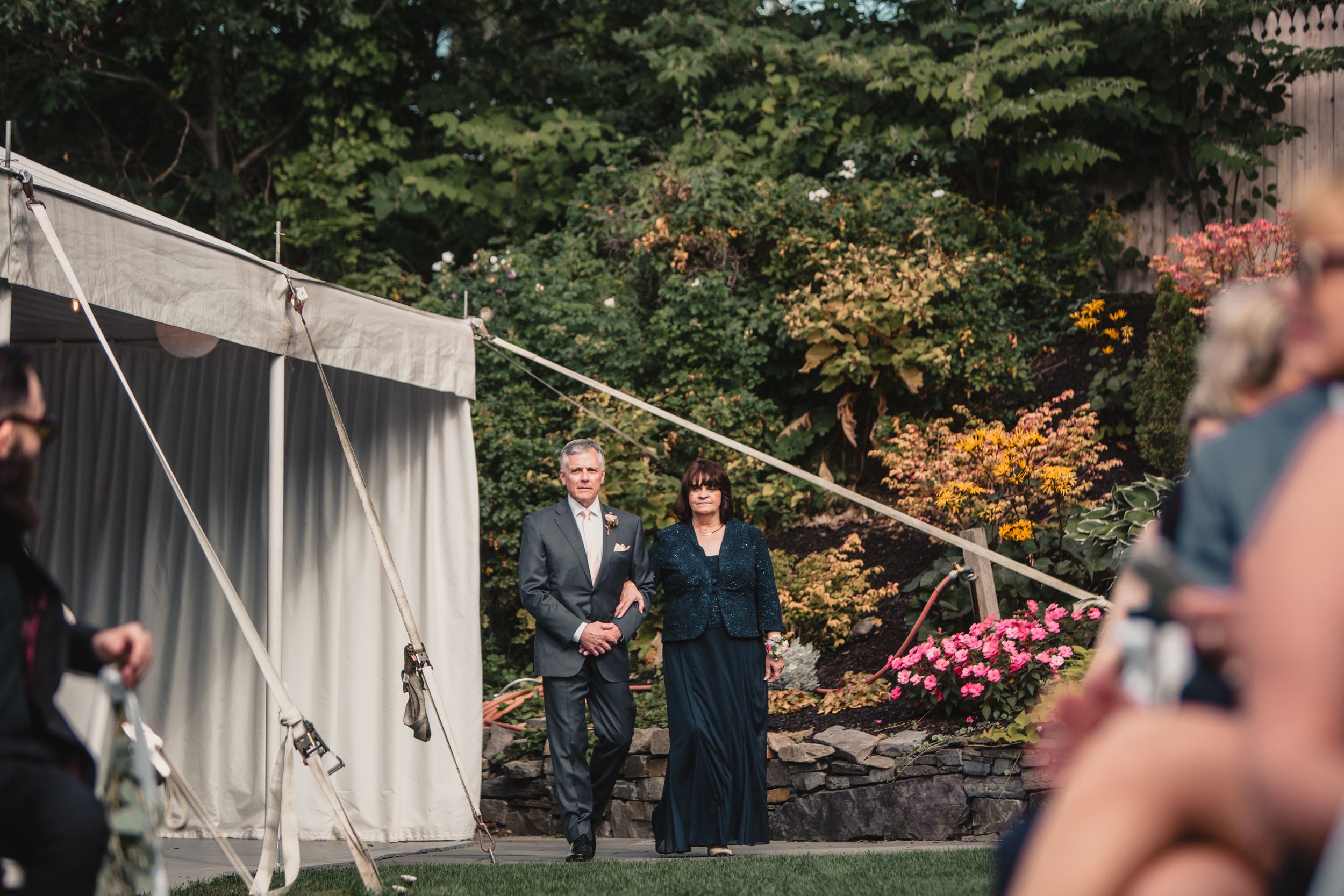 Couple walking together outdoors at a wedding venue with a white tent and lush green gardens in the background.