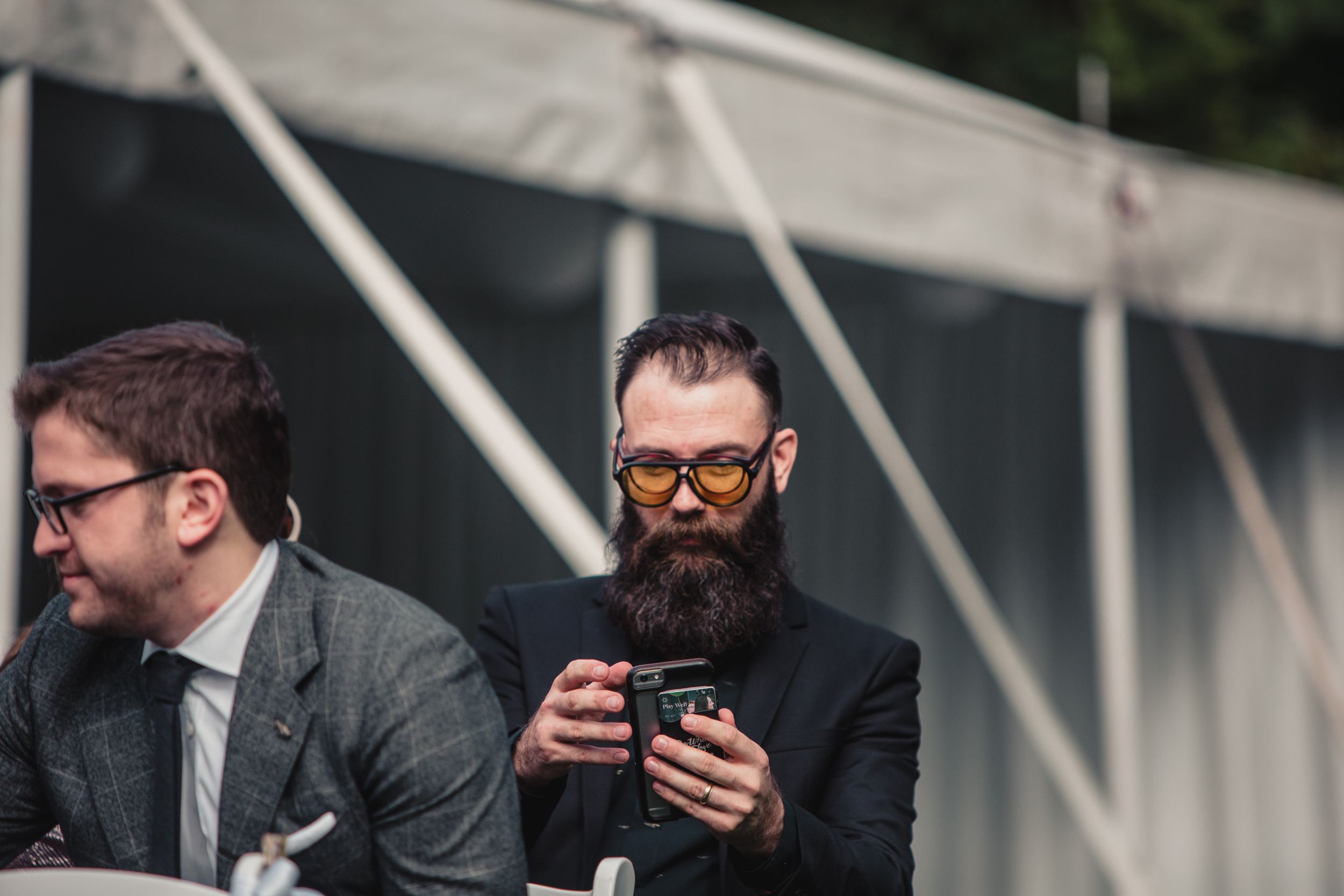 Two stylish men in suits sit outdoors, one with a long beard and amber sunglasses using a smartphone.