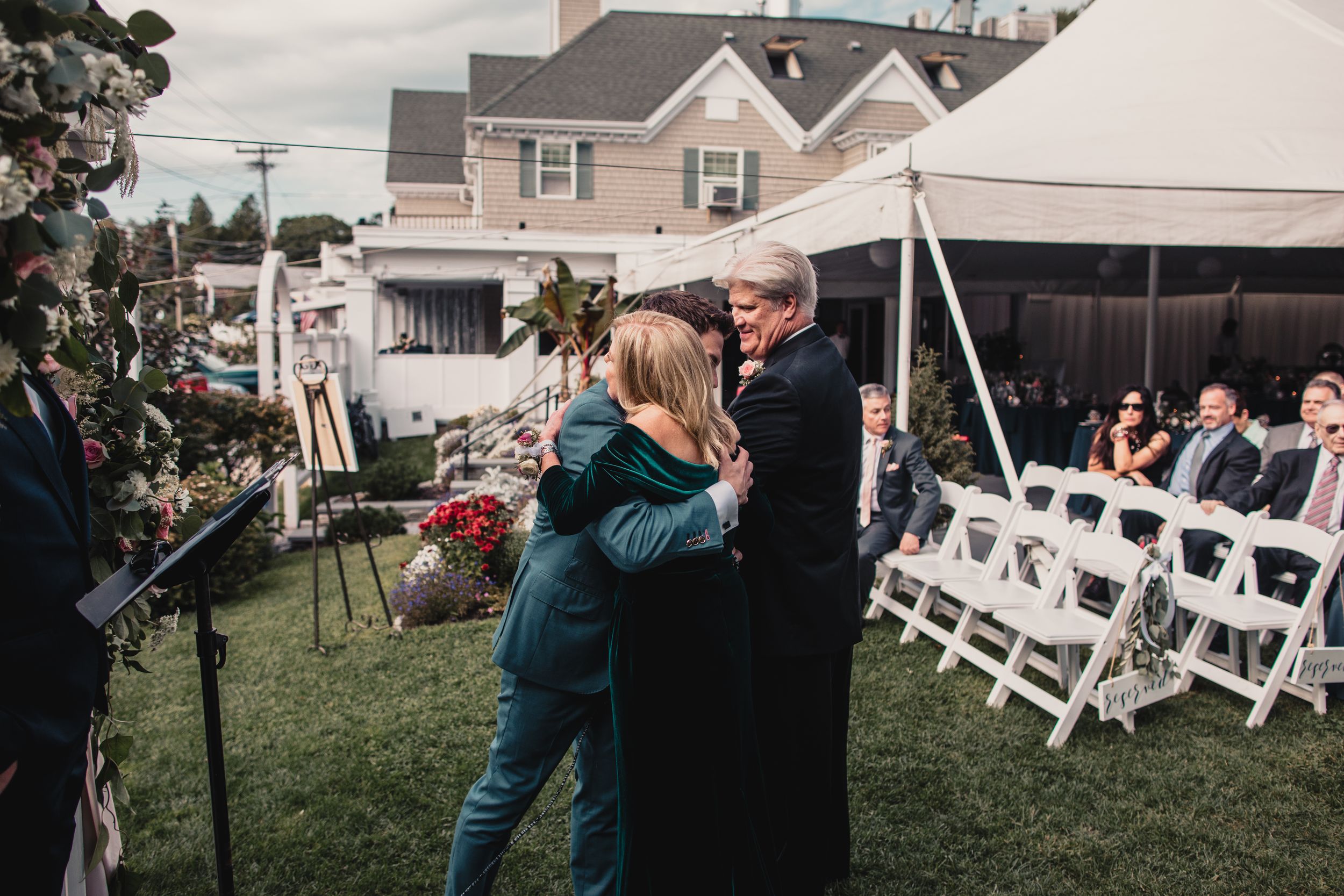 Outdoor wedding ceremony with guests seated in white chairs as couple embraces near floral arrangements.