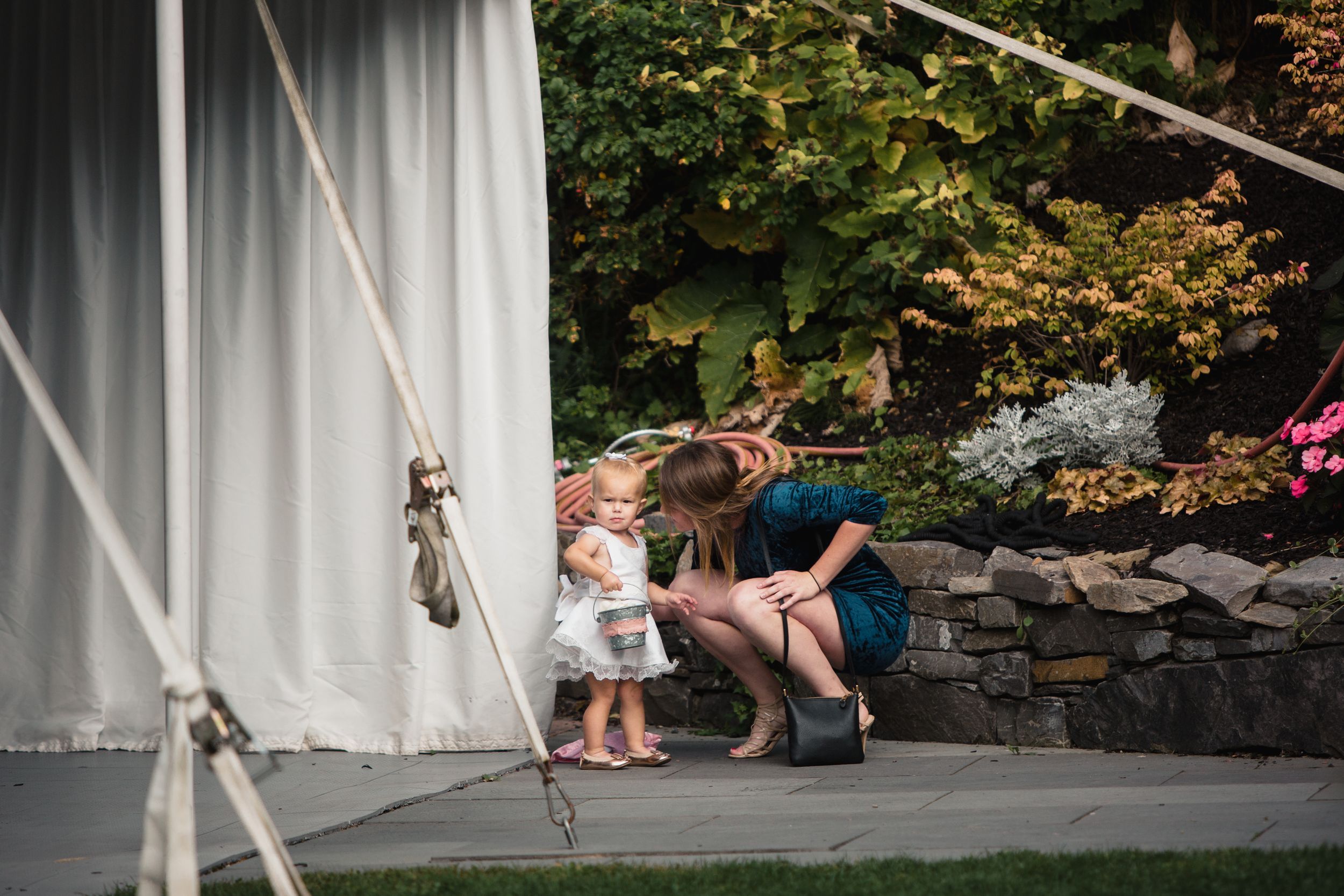 Woman crouching to interact with a toddler girl near a white tent at an outdoor garden event.