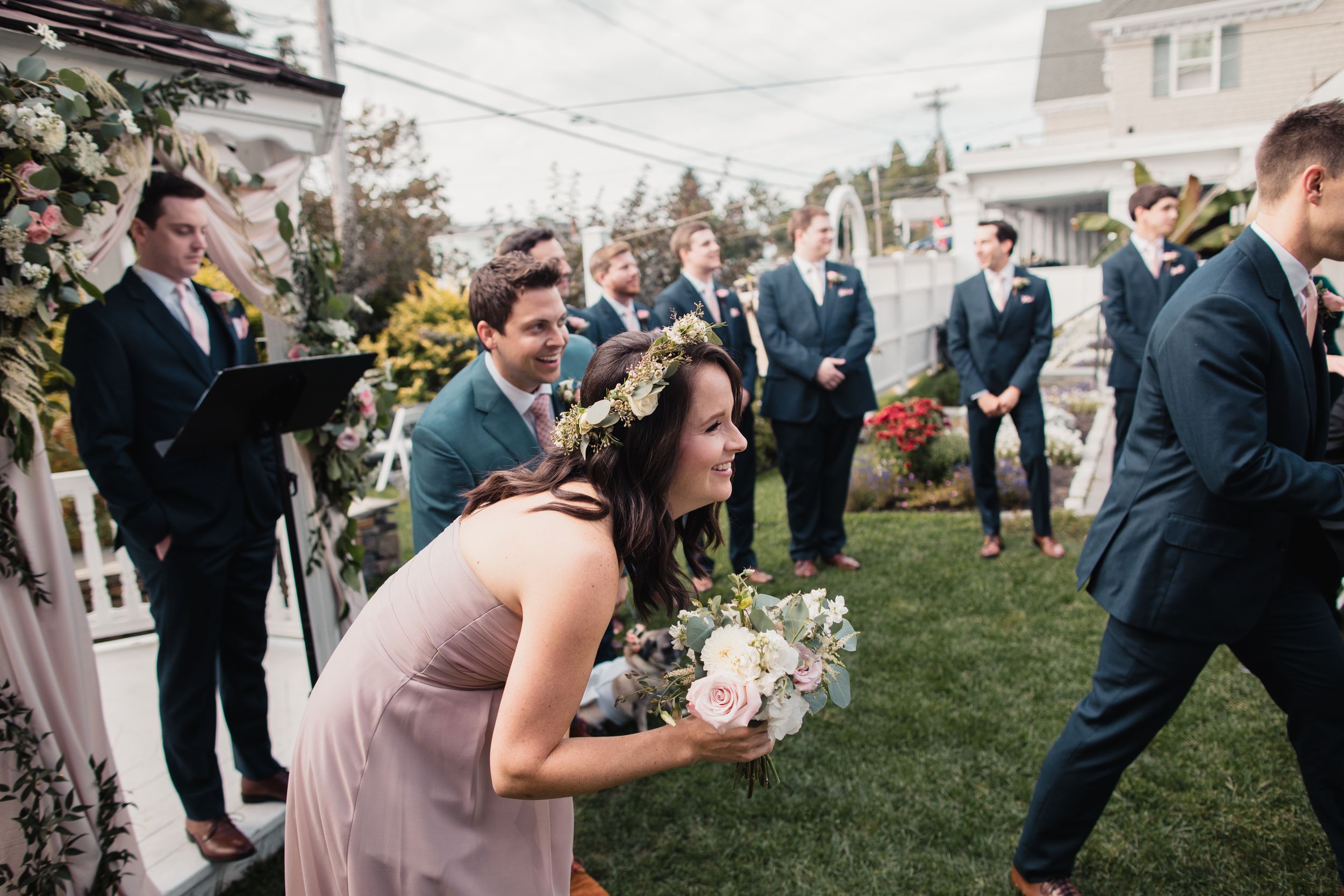 Bridesmaid in blush dress kneeling on grass during outdoor wedding ceremony with groomsmen in navy suits.