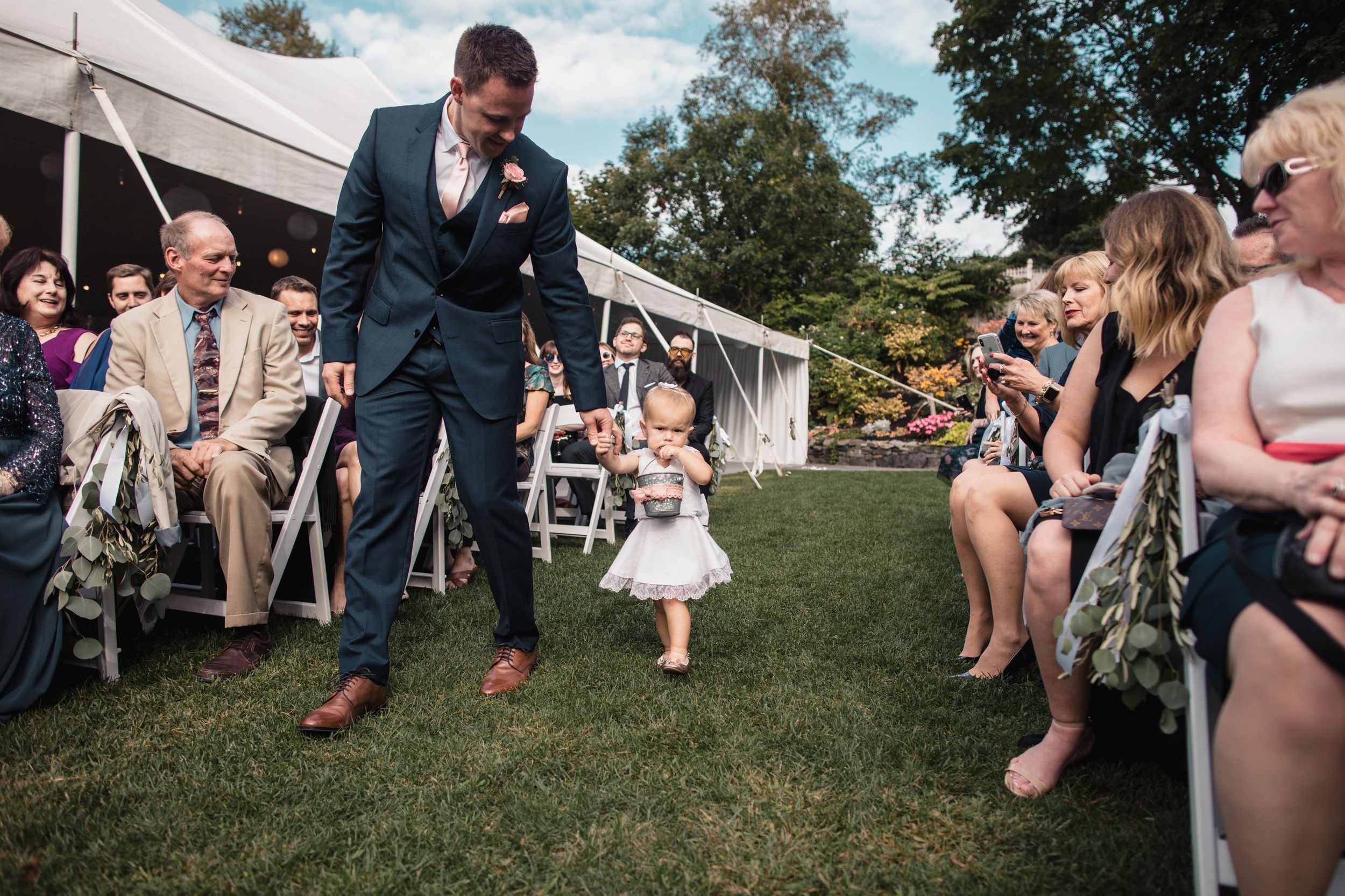 Groom walks baby flower girl down the aisle at outdoor wedding ceremony while guests watch.