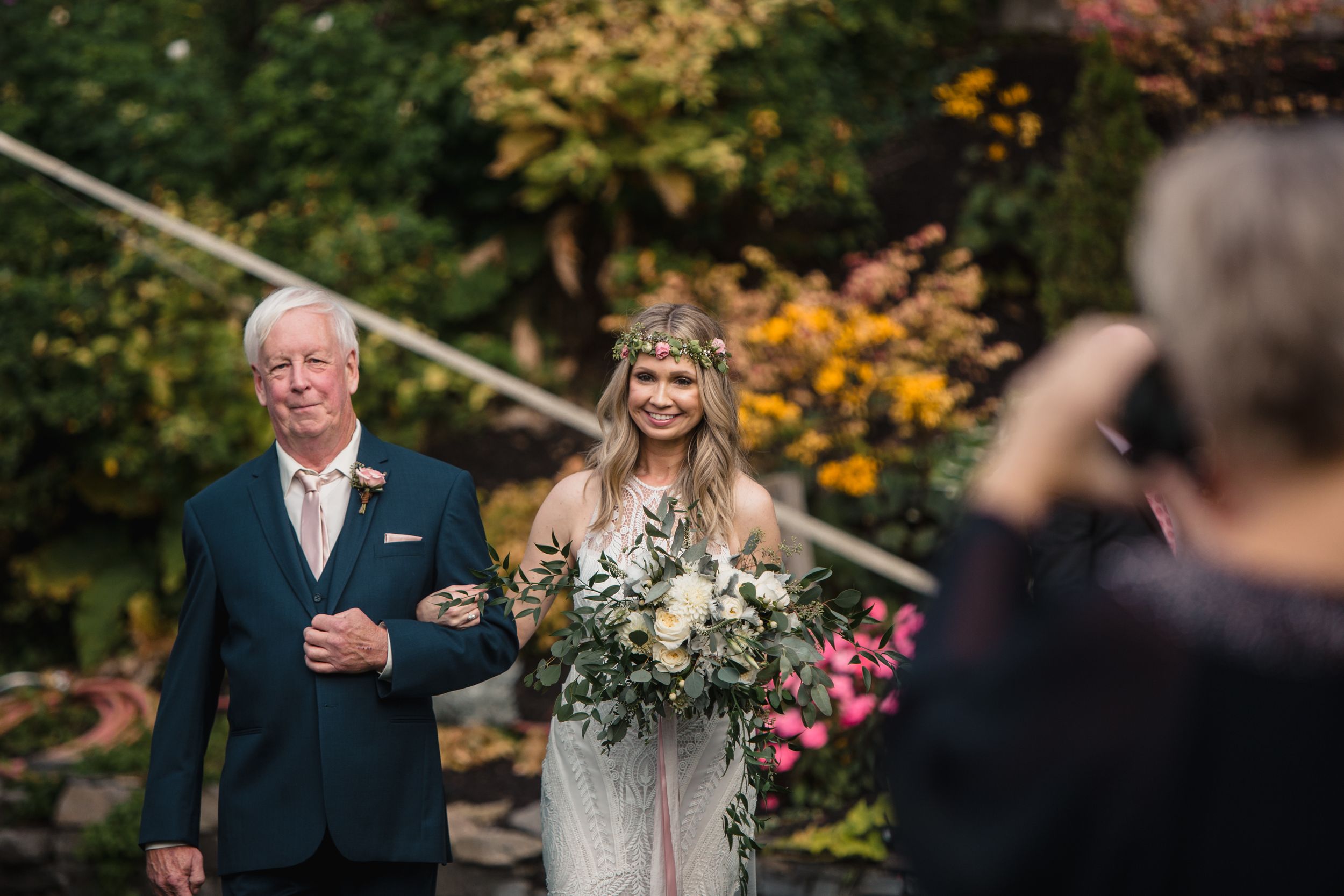 Bride in floral crown holding bouquet walks with older man in garden wedding ceremony.