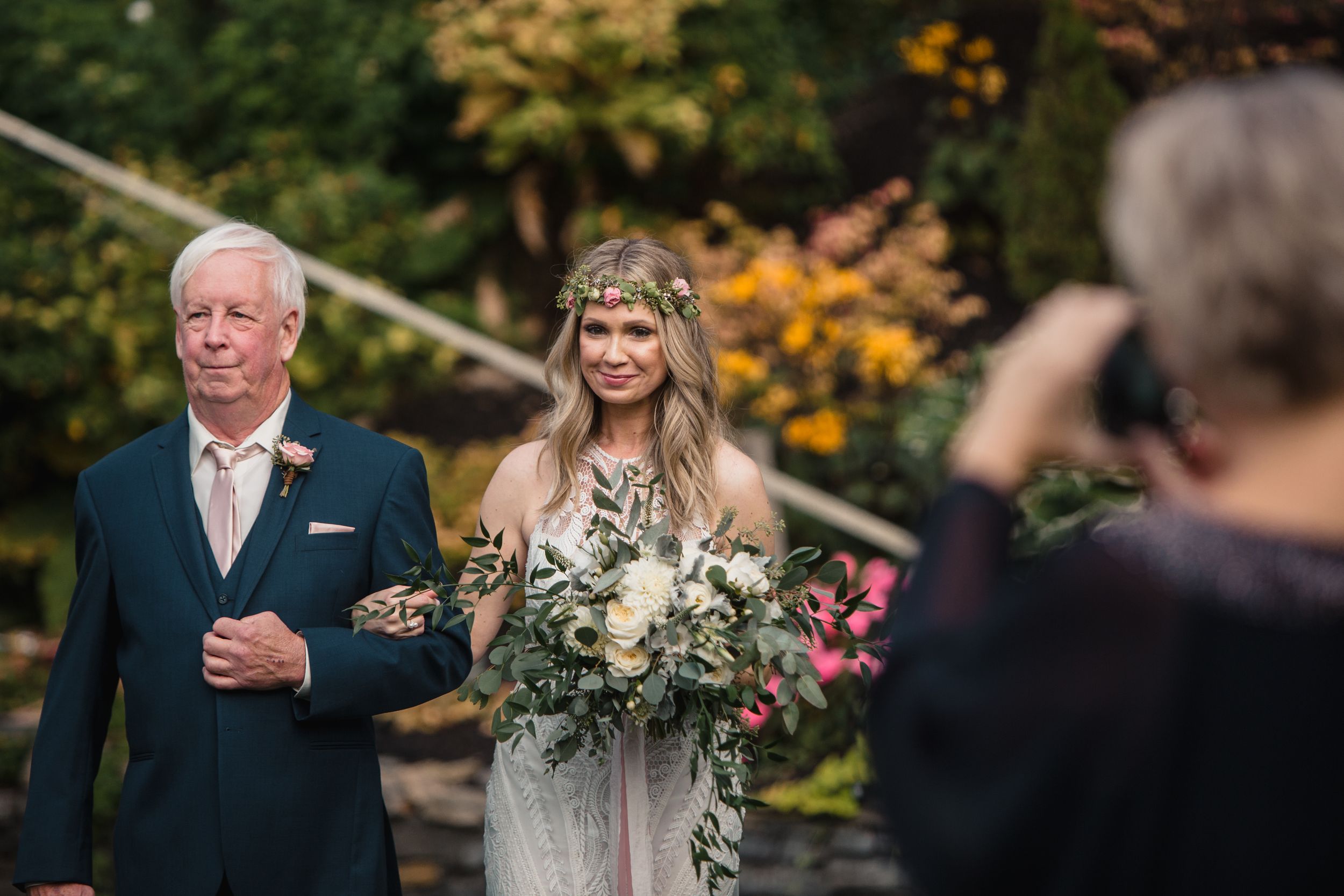 Bride in floral crown holding bouquet walks with older man at outdoor garden wedding ceremony.