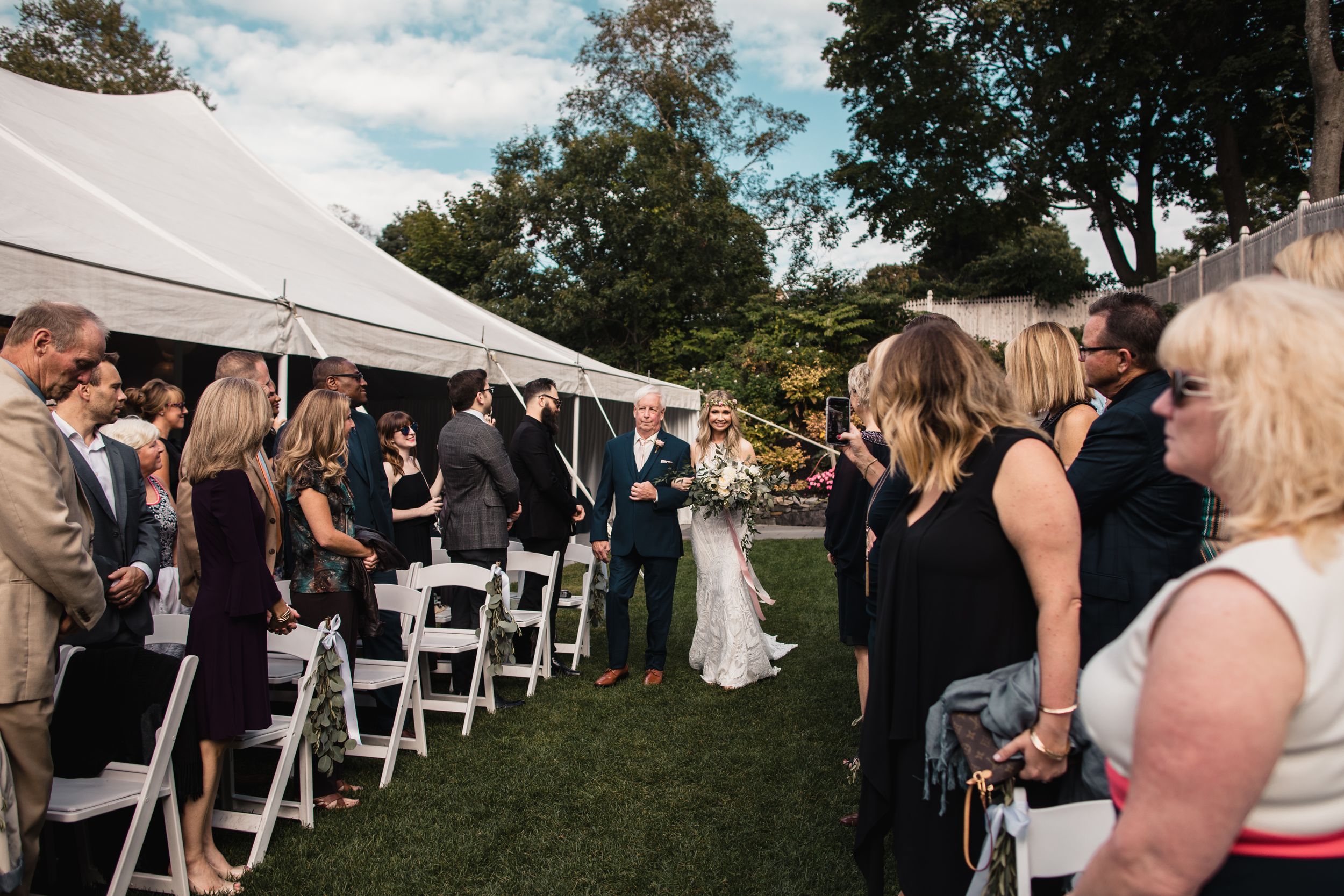Bride walks down outdoor wedding aisle flanked by guests in formal attire, with white tent and lush greenery backdrop.