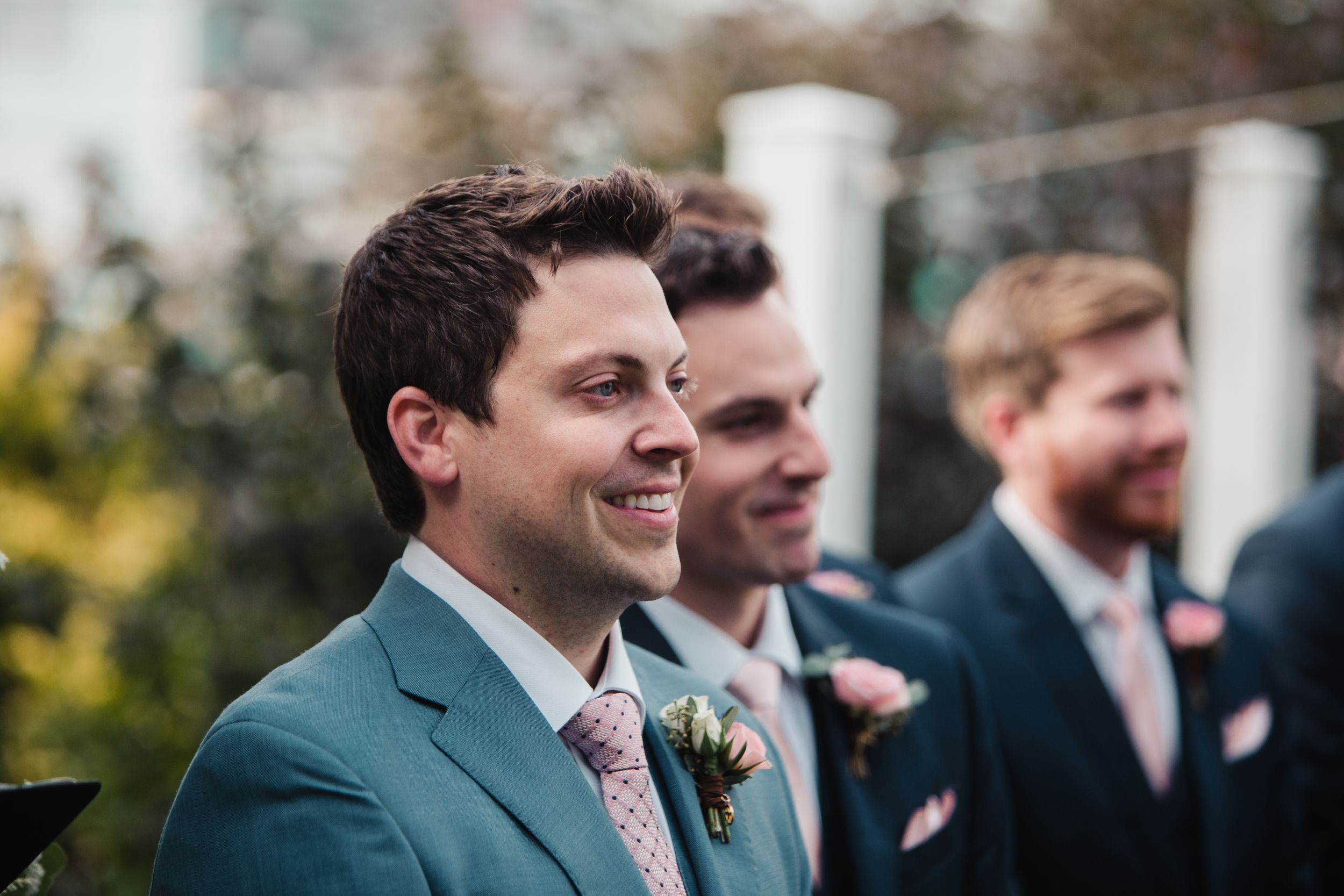 Smiling groom in teal suit with boutonniere stands with groomsmen in navy suits at outdoor wedding.