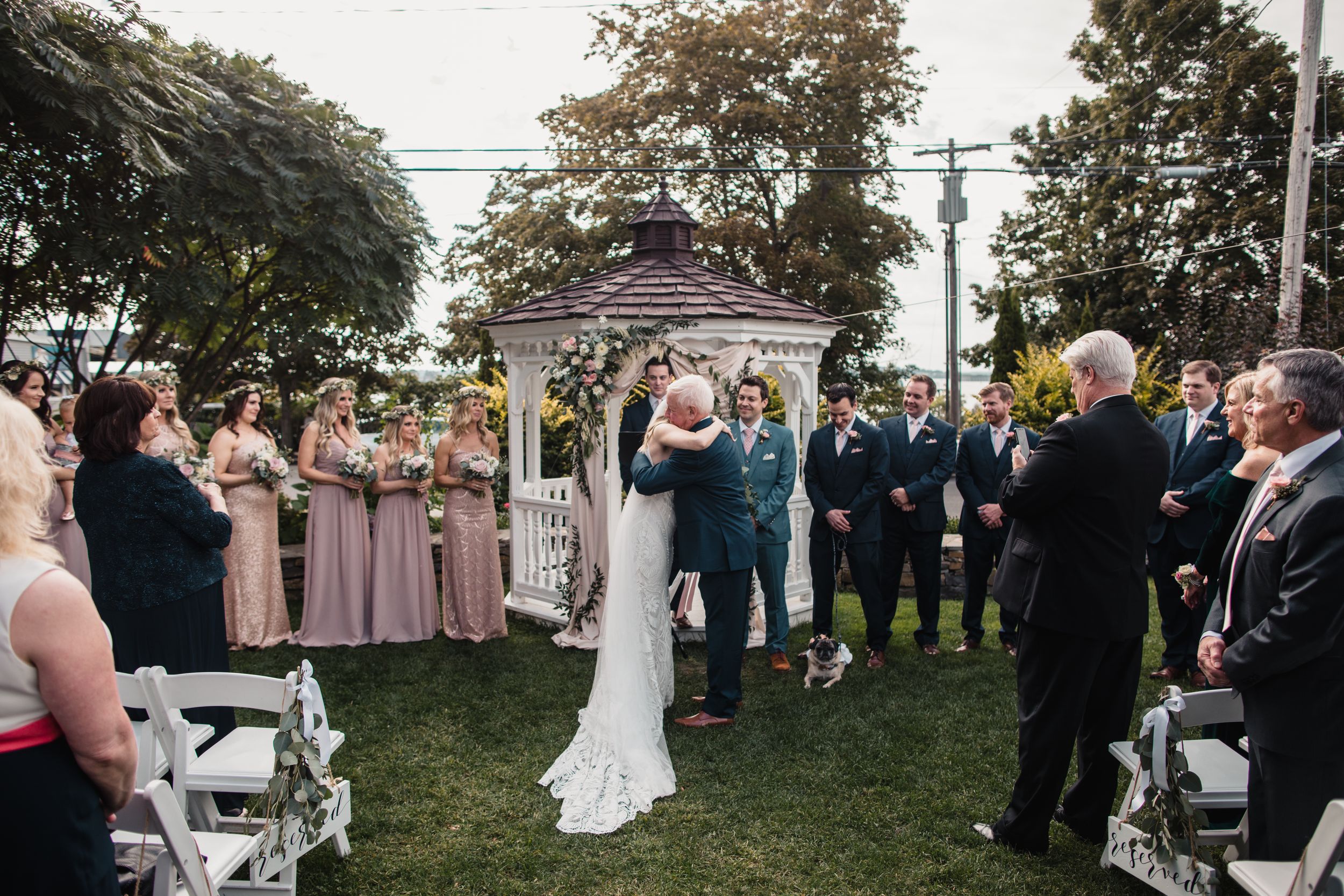Outdoor wedding ceremony with bride and groom kissing near a white gazebo, bridesmaids in pink dresses and groomsmen in blue suits.