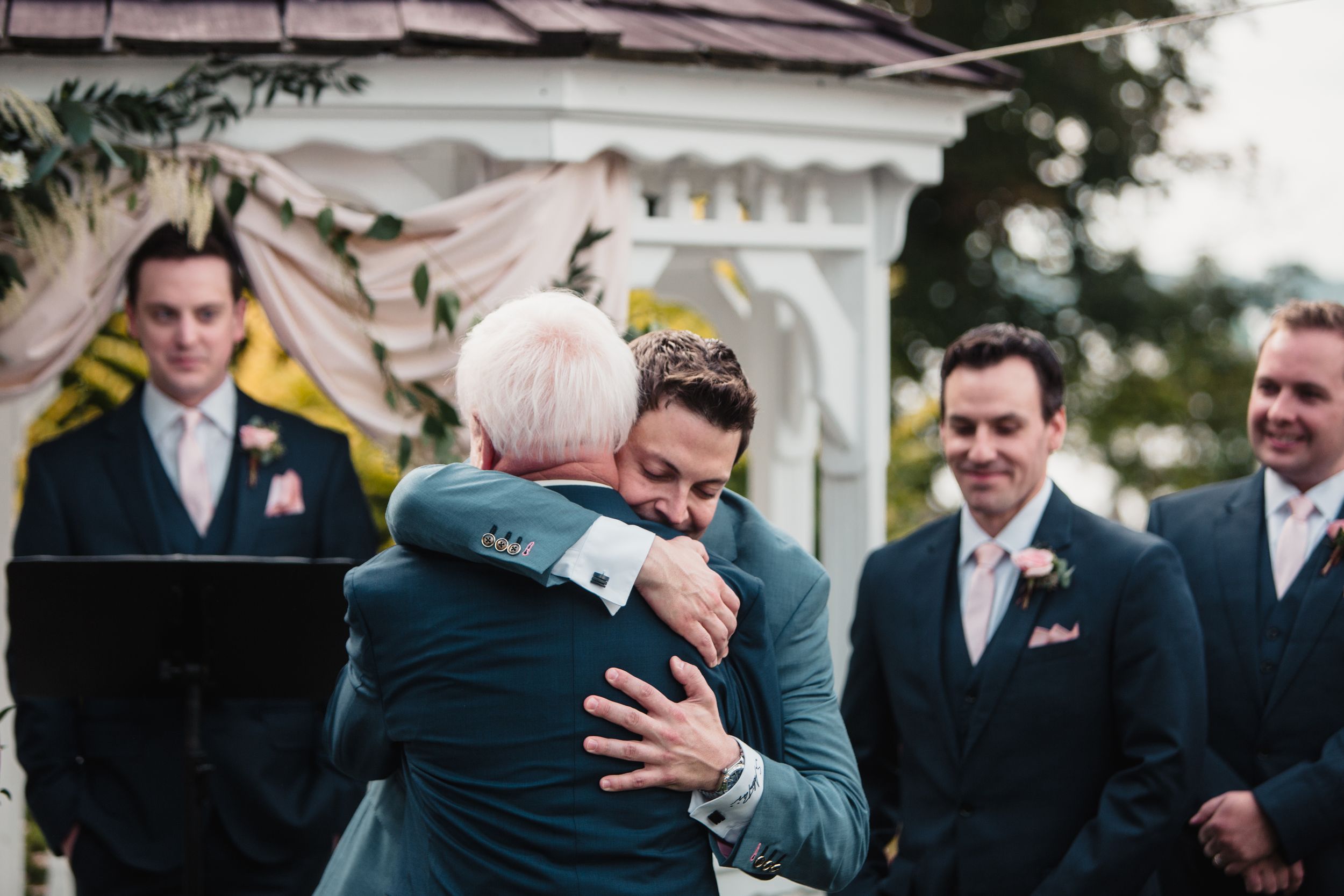 Groom in teal suit shares emotional hug with older man at outdoor wedding ceremony gazebo.