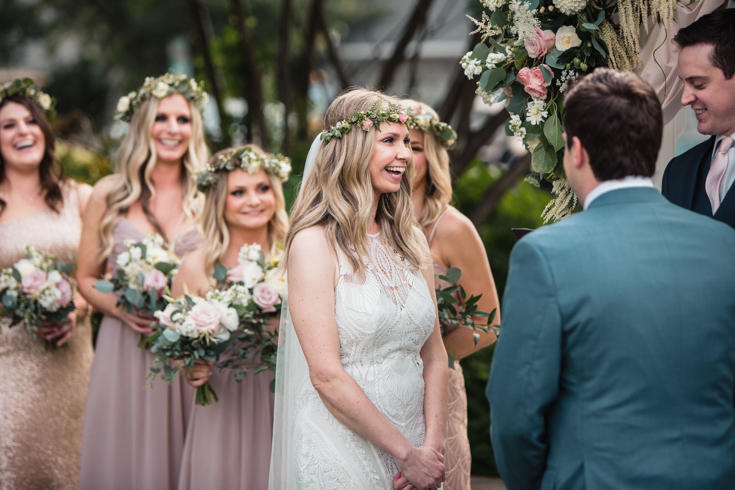Smiling bride wearing a floral crown stands with bridesmaids holding white bouquets during an outdoor wedding ceremony.