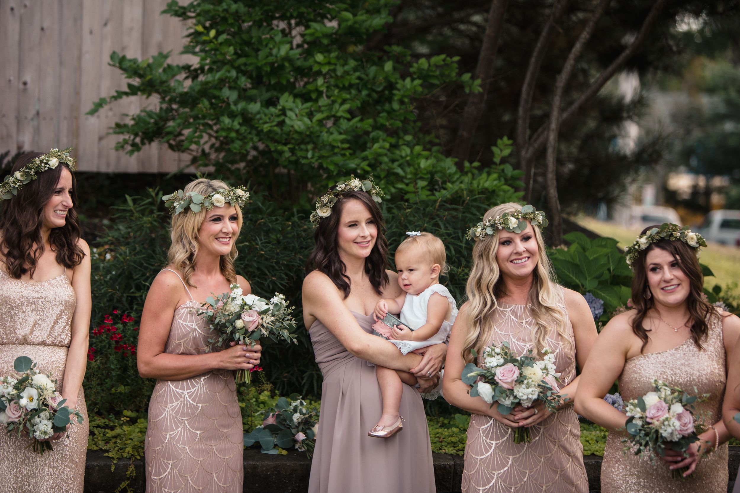 Five bridesmaids in champagne dresses with floral crowns hold bouquets, one holding a baby.