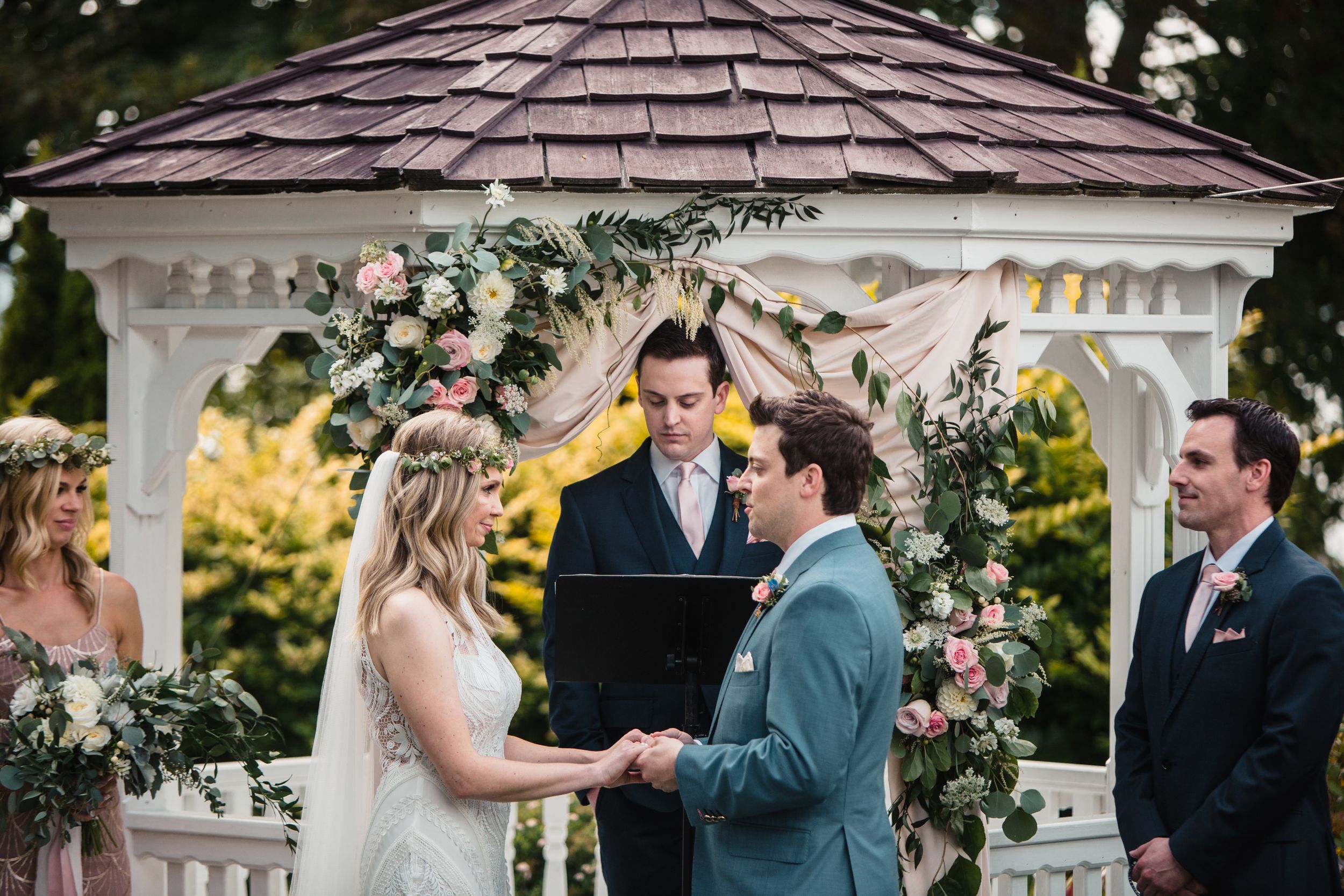 Bride and groom exchange vows under floral-adorned gazebo at outdoor wedding ceremony.