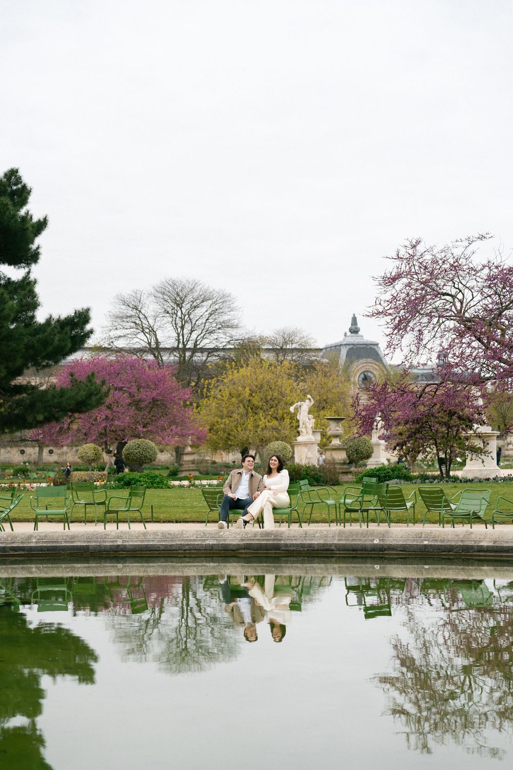 Couple sitting by a reflective pond in Tuileries Garden, Paris, with blooming trees. Cherry blossoms editorial engagement photoshoot