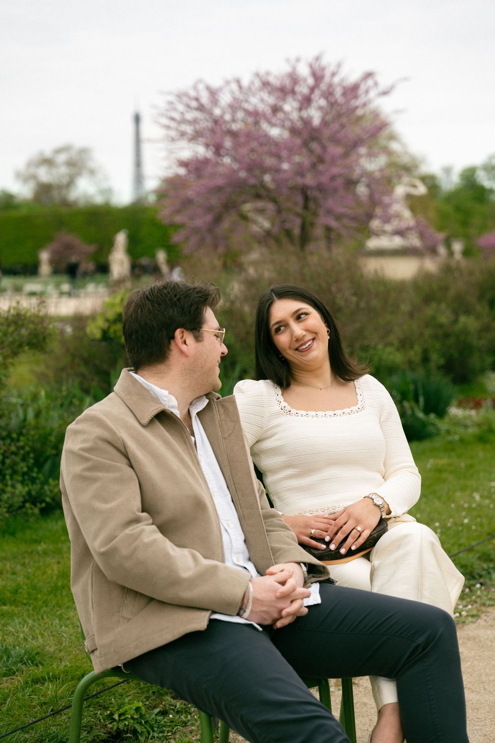 Couple sitting on a bench in a Paris garden with the Eiffel Tower and pink blossoms in background. Paris editorial wedding photographer