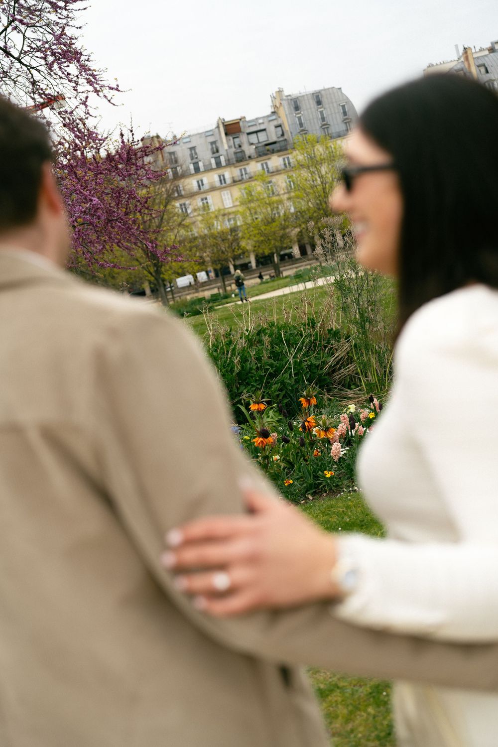 Couple embracing in Parisian garden with colorful tulips and classic Haussmann buildings. Paris editorial engagement photographer