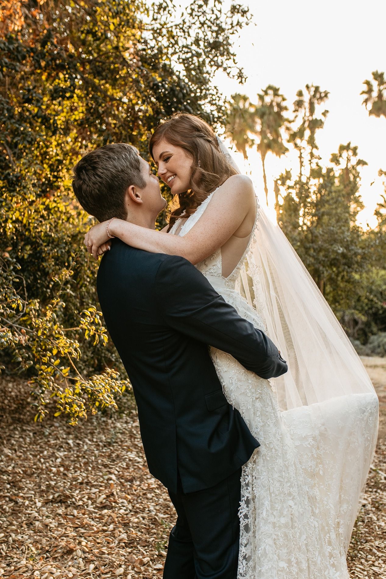Groom lifting bride in white gown and veil, sharing a romantic moment outdoors with palm trees at golden hour at Rancho Buena Vista Adobe.