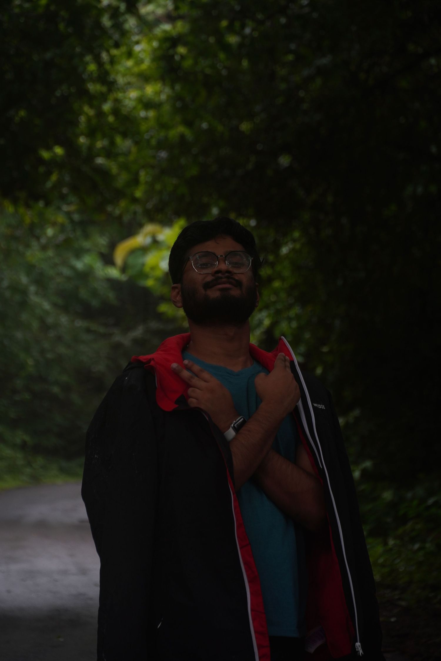 A moody, vertical portrait of a trekker in a red jacket walking through the deep shadows of an ancient forest trail.