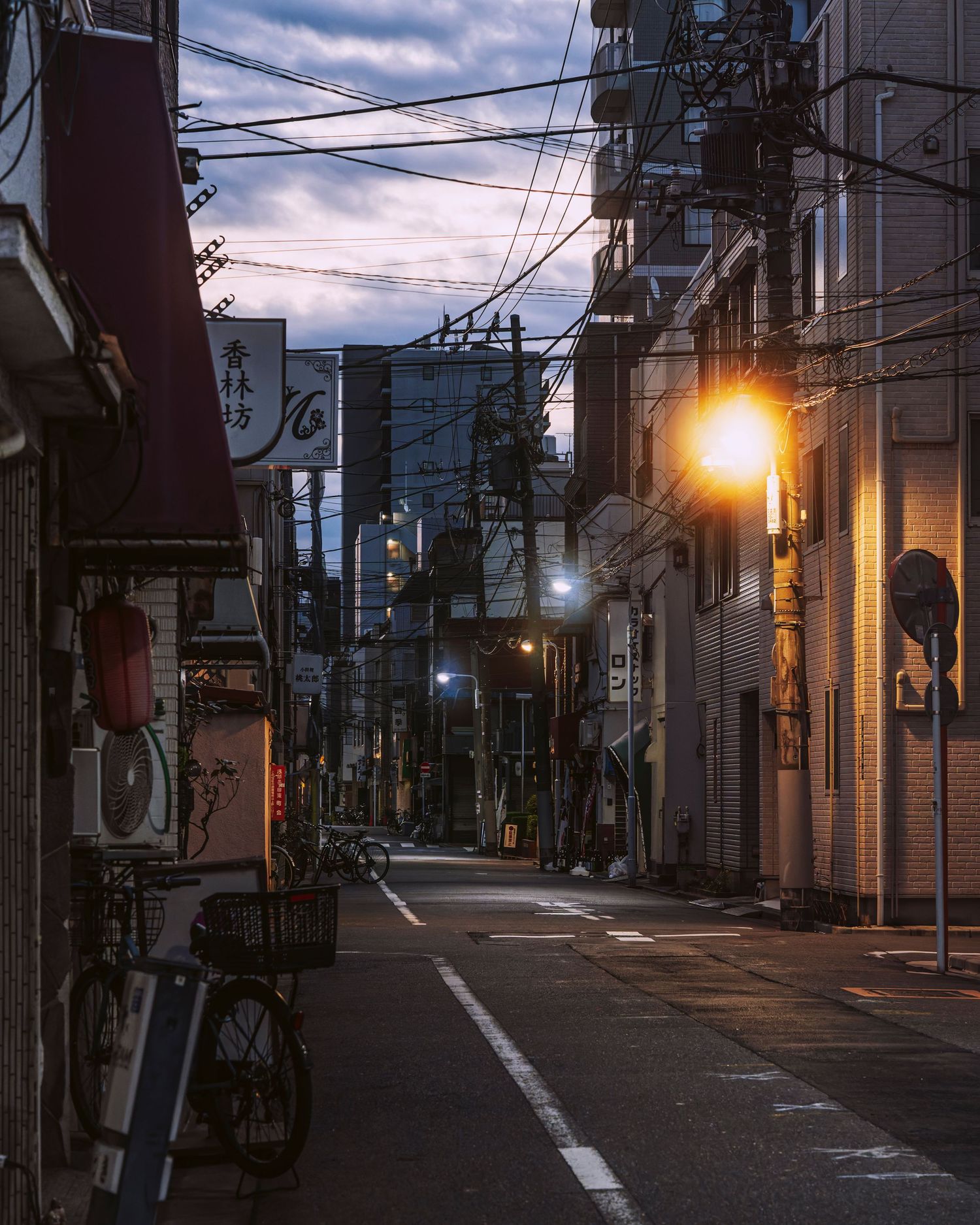 A quiet Japanese alley at dusk with glowing street lights, utility poles, and traditional shop signs lining the narrow road.