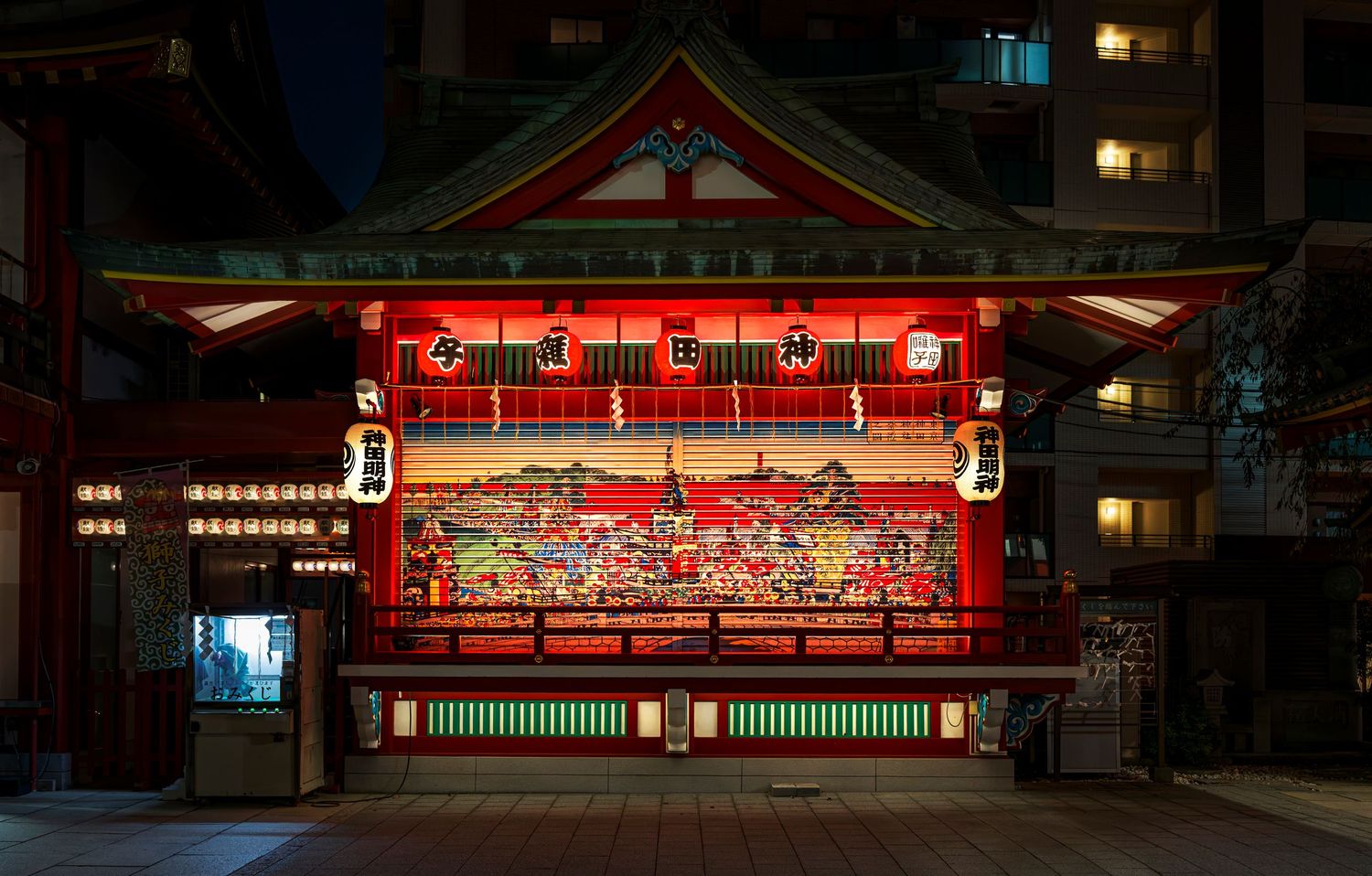Illuminated Japanese shrine with vibrant red facade and traditional artwork glowing at night in urban setting.