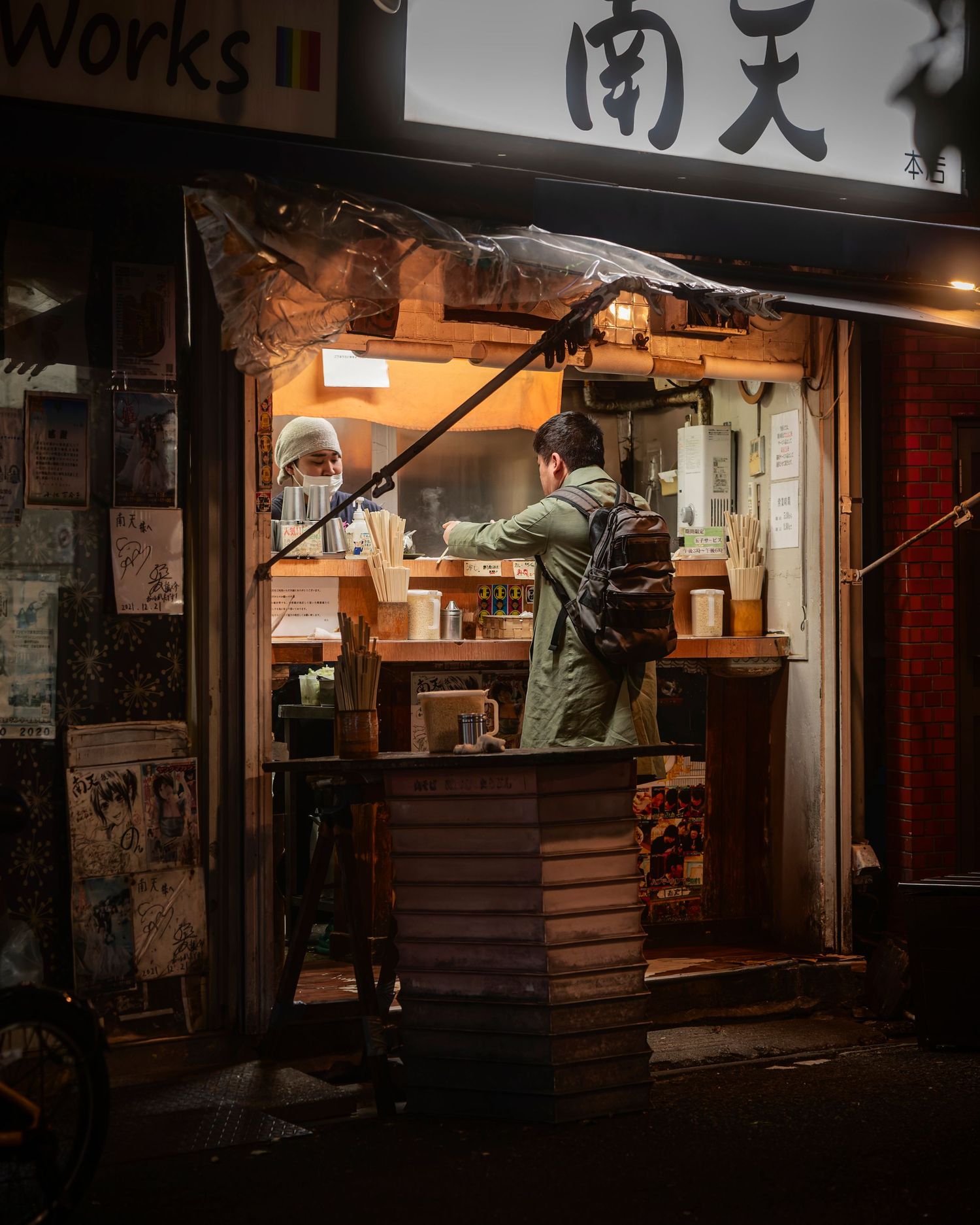 Person ordering at a small Japanese street food stall at night, with warm glowing lights and kanji signage.